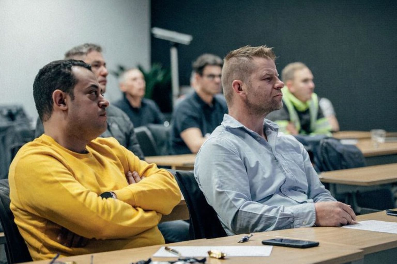 Menn sitter i et møterom, lytter oppmerksomt til en presentasjon.