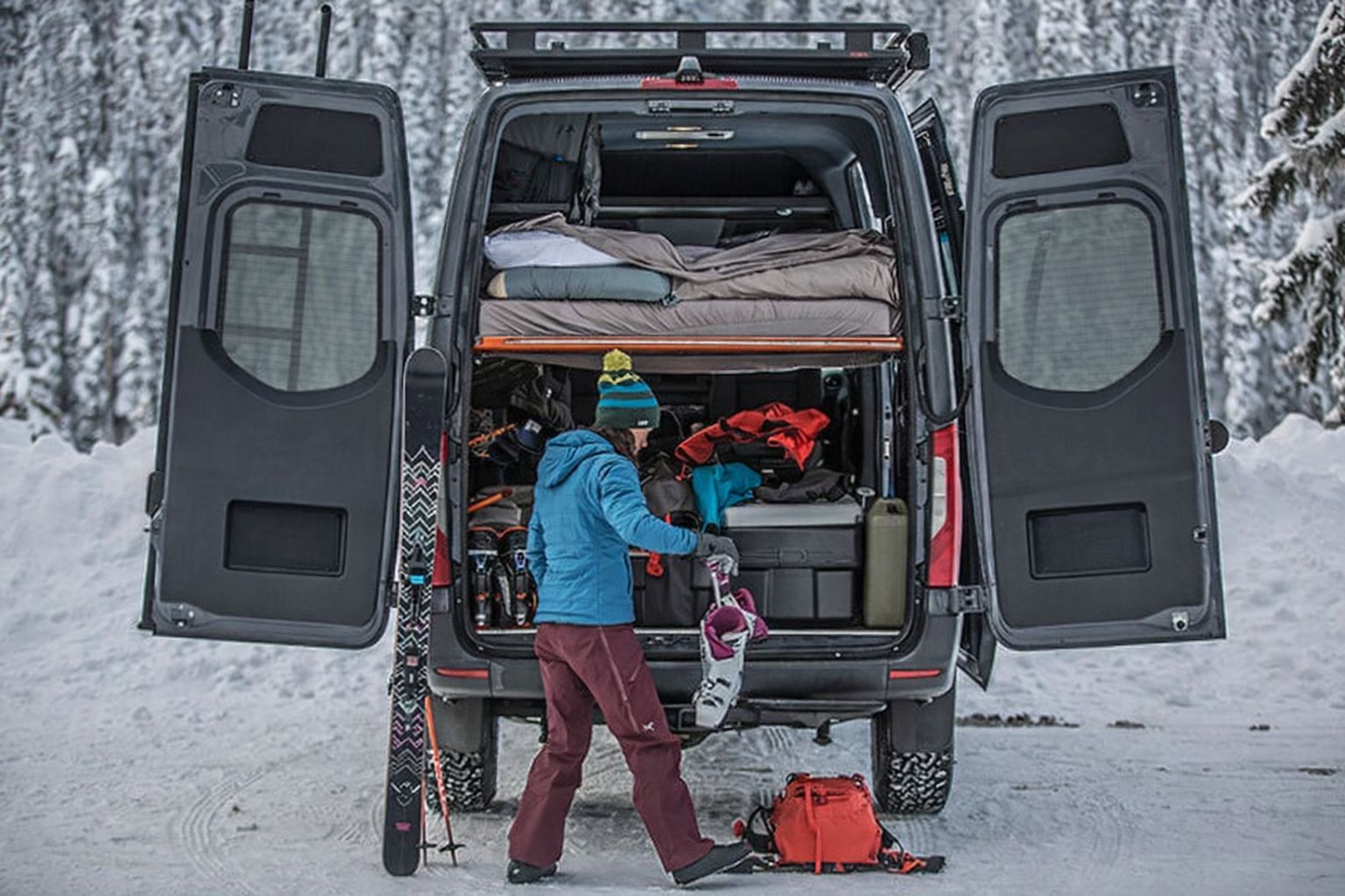Varebil med åpen bakluke, snø, og en person som forbereder seg på ski.